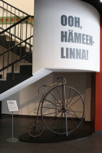 Vanhanaikainen polkupyörä. Suuri eturengas ja paljon pienempi takarengas. Satula eturenkaan päällä. Pyörän yläpuolella seinässä teksti "Ooh, Hämeenlinna!".