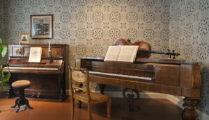 Vasemmalla piano, oikealla taffelipiano. Soittimien edessä tuolit, nuottitelineillä nuotteja. Taffelipianon päällä sello.