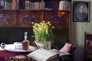 Palanderin talon herrainhuone. Pöydällä on narsisseja ruukussa, konjakkipullo ja neljä lasia hopeatarjottimella sekä avonainen raamattu.