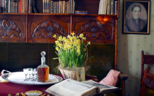 Palanderin talon herrainhuone. Pöydällä narsisseja korissa, avoin Raamattu sekä tarjottimella konjakkipullo ja neljä lasia.