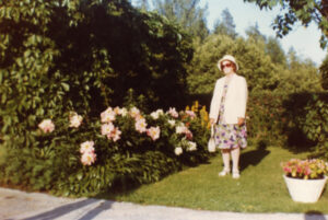 Nainen seisoo vehreässä puutarhassa alppiruusupensaan vieressä. Värivalokuva.