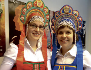 Kaksi naista värikkäissä asuissa ja päähineissä. Toisen päähine punainen, toisen sininen. Molemmissa paljon koristeita ja helmiä.