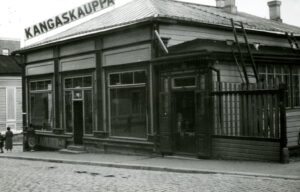 Puutalo, jonka katolla Kangaskauppa-kyltti. Kolme näyteikkunaa, yhden edessä ihminen. Mustavalkovalokuva.