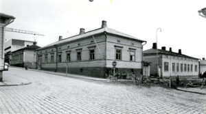 Neljän tien risteys. Teiden varsilla matalia puu- ja kivitaloja. Oikealle lähtevä tie suljettu tietyön vuoksi. Näkyvissä kaksi työntekijää. Mustavalkovalokuva.