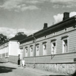 Kadun varressa yksikerroksisia puurakennuksia. Etummaisen seinässä kyltti, jossa lukee Firestone sekä nuoli jossa lukee "Autokorjaus". Mustavalkovalokuva.