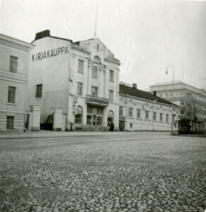 Etualalla Hämeenlinnan toria. Torin laidalla suuri kolmikerroksinen kivirakennus. Sen päädyssä ja oven yläpuolella lukee Kirjakauppa. Oikealla puolella on vaalea, yksikerroksinen puurakennus. Mustavalkovalokuva.