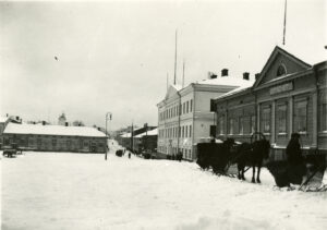 Vasemmalla lumista Hämeenlinnan toria. Oikealla torin laidan rakennuksia. Etummaisena yksikerroksinen puurakennus, jonka seinässä kyltti Kaupungin hotelli. Rakennuksen edessä kolmet hevoskärryt ajureineen. Mustavalkovalokuva.