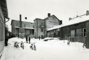 Etualalla luminen piha. Pihassa ihmisiä ja muutama polkupyörä lumessa. Takana kaksikerroksinen rakennus, osin puuta ja osin kiveä. Oikealla matalampi, tumma puurakennus. Mustavalkovalokuva.