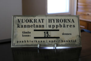 Valkoinen kyltti, jossa mustalla teksti "Vuokrat kannetaan/Hyrorna uppbäres tämän kuun/dennes 15. pankkiaikana/under banktid".
