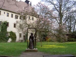 Kuvan keskellä pronssipatsas naisesta pitkässä mekossa. Ympärillä nurmikkoa. Takana puu, jossa vaaleanpunaisia kukkia, sekä vaalea suuri kivirakennus.