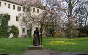 Kuvan keskellä pronssipatsas naisesta pitkässä mekossa. Ympärillä nurmikkoa. Takana puu, jossa vaaleanpunaisia kukkia, sekä vaalea suuri kivirakennus.