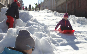 Lapsi laskee punaisella pulkalla alas lumista mäkeä. Taustalla ja vasemmassa reunassa muita ihmisiä.