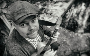 Mies, jolla on hattu päässä ja ukulele käsissä. Yläviistosta otettu mustavalkovalokuva.