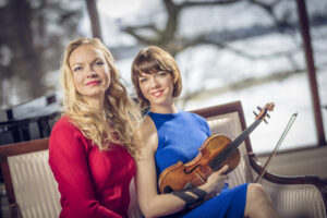 Kaksi naista istuu penkillä. Toisella punainen ja toisella sininen asu. Siniasuisella, oikeanpuoleisella naisella viulu.
