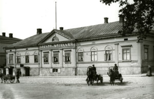 Vaalea suuri puurakennus korkealla kivijalalla. Seinässä teksti "Stads hotellet". Edessä kahdet hevoskärryt kuskeineen. Mustavalkovalokuva.