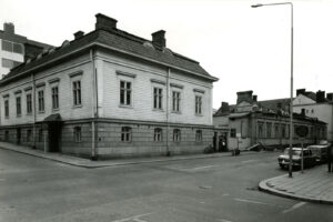 Kadun kulmassa suurehko puurakennus korkealla kivijalalla. Etualalla asfalttitie, jonka oikeassa laidassa kaksi pysäköityä autoa. Mustavalkovalokuva.