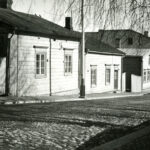 Yksi- ja kaksikerroksisia puurakennuksia mukulakivikadun vasemmalla laidalla. Katu etualalla.. Mustavalkovalokuva.