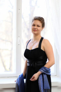 Nainen seisoo ikkunan edessä. Yllä musta mekko, käsivarsilla sininen huivi.