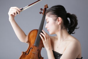 Nuori tummahiuksinen nainen pitelee viulua silmät kiinni. Toinen käsi kohotettu ylös, ja siinä viulun jousi.