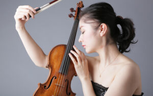 Nuori tummahiuksinen nainen pitelee viulua silmät kiinni. Toinen käsi kohotettu ylös, ja siinä viulun jousi.