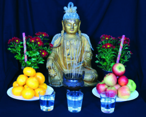 Keskellä kulanvärinen Buddha-patsas. Patsaan molemmin puolin kukkia. Edessä omenia ja appelsiineja lautasilla sekä kolme vesilasia.