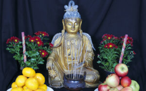 Kullanvärinen Buddha-patsas. Patsaan molemmilla puolilla kukkia. Edessä omenia ja appelsiineja lautasilla.