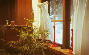 Oikealla ikkuna, jossa valkoiset verhot. Ikkunalaudalla kaksi kynttilää punaisissa jaloissa. Ikkunan edessä pieni kuusi. Taustalla ruskea kaappi.