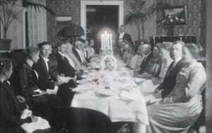 Ihmisiä istumassa pitkän pöydän ääressä juhlavaatteet päällä. Pöydän keskellä kynttilöitä. Mustavalkovalokuva.