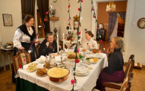 Neljä 1800-luvun lopun asuihin pukeutunutta naista ruokapöydän ympärillä. Yksi seisoo, muut istuvat. Pöydässä on jouluruokia ja pöydän yläpuolelta roikkuvat värikkäät joulukoristeet.