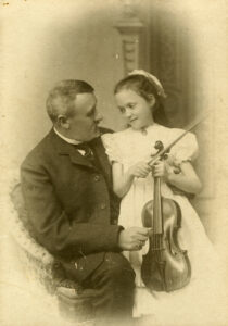Mies istuu tuolilla, tyttö polvellaan. Tytöllä vaalea mekko ja käsissään viulu. Miehellä tumma puku. Katsovat toisiaan. Mustavalkovalokuva.
