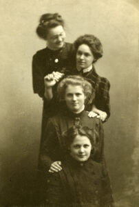Neljä naista poseeraa. Pitkähihaiset, korkeakauluksiset puvut päällä. Mustavalkovalokuva.
