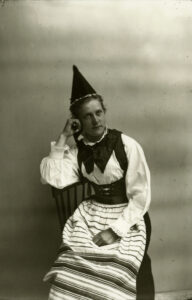 Tuolilla istuva nainen, jolla kansallispuvun tyylinen asu ja päässä suippo hattu. Mustavalkovalokuva.