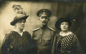 Kolme henkilöä. Keskellä viiksekäs mies sotilasasussa, koppalakki päässä. Reunoilla kaksi naista, joilla koristeelliset hatut. Mustavalkovalokuva.