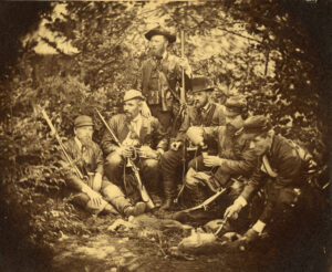 Kuusi miestä poseeraa metsässä aseet käsissä. Oikeanpuolimmainen nylkee maassa olevaa jänistä. Mustavalkovalokuva.