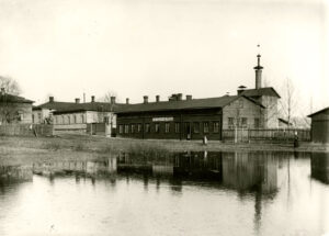 Etualalla vettä, taustalla asuinrakennuksen näköinen pitkä, matala puurakennus. Rakennuksen seinässä teksti C. O. Saxelin oluttehdas. Mustavalkovalokuva.