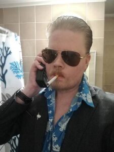 Mies, jolla on yllä tumma takki ja sininen paita. Suuret tummat aurinkolasit päässä, vanhan mallinen matkapuhelin korvalla ja tupakka suussa.
