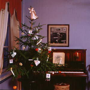 Joulukuusi Sibeliuksen syntymäkodissa. Kuusi on koristeltu 1800-luvun lopun tyylisillä, käsin tehdyillä paperikoristeilla ja kynttilöillä. Taustalla piano.