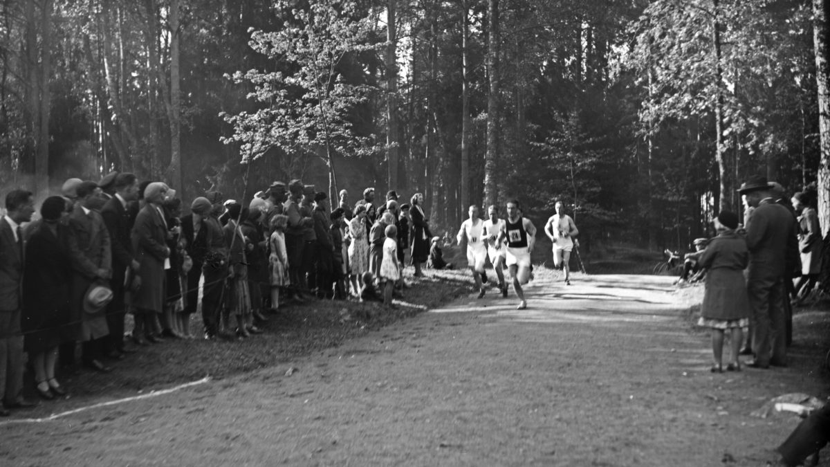 Ihmiset juoksevat valkoisissa shortsipuvuissa metsäisellä hiekkatiellä auringonpaisteessa. Tien molemmin puolin seisoo paljon yleisöä. 