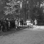 Ihmiset juoksevat valkoisissa shortsipuvuissa metsäisellä hiekkatiellä auringonpaisteessa. Tien molemmin puolin seisoo paljon yleisöä.