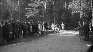Ihmiset juoksevat valkoisissa shortsipuvuissa metsäisellä hiekkatiellä auringonpaisteessa. Tien molemmin puolin seisoo paljon yleisöä.
