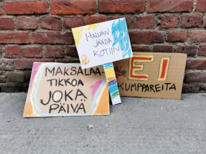 Hippaloiden mielenosoituskylttityöpajan mainoskuva: Kolme pahvista tehtyä värikästä mielenosoituskylttiä, joissa lukee "Maksalaatikkoa joka päivä", "Haluan jäädä kotiin" ja "Ei kumppareita".