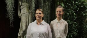 Kaksi vaaleapaitaista naista hymyilee kameralle. Tummassa taustassa köynnöskasveja ja osa kreikkailaista patsasta.