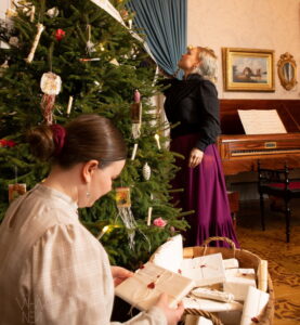 Kaksi naista valmistelee joulua Palanderin talon salissa. Toinen järjestelee puotipaperiin käärittyjä paketteja ja toinen koristelee tunnelmallista joulukuusta.