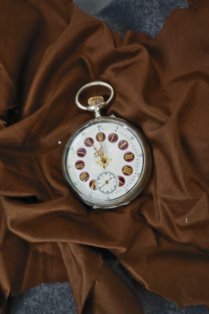 Taskukellon muotoinen hopeinen kello aseteltuna ruskean kankaan päälle. Kellossa on valkealla taustalla punaiset ympyrät roomalaisten numeroiden ympärillä sekä koristeelliset kullanväriset viisarit. Kellotaulussa on lisäksi alaosassa pienempi taulu.