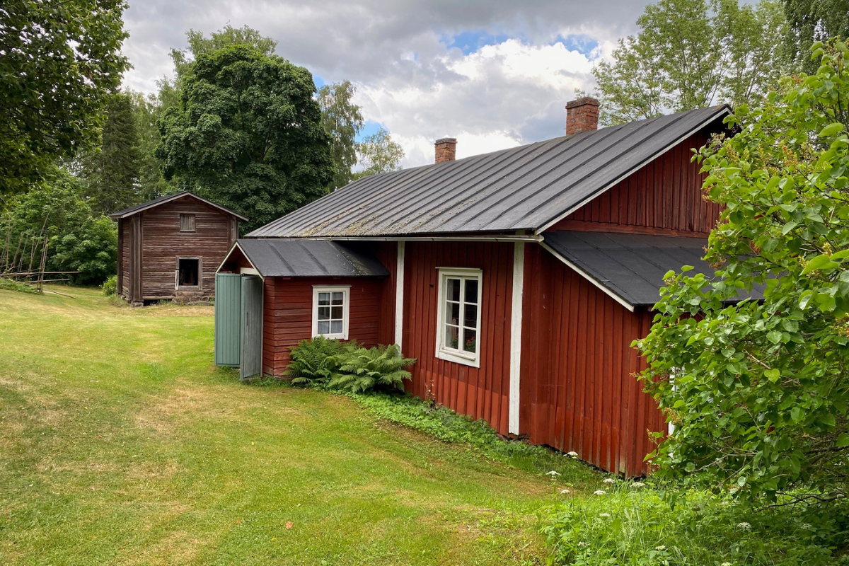 Kuvassa vanha punainen puinen rakennus ulkoa kesällä vasta leikatun nurmikon oikealla puolella.