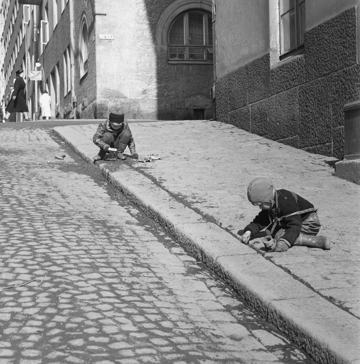 Näyttelyssä esilläoleva kuva, jossa kaksi lasta leikkii mukulakivikadulla maassa istuen. 