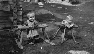 Kaksi pientä lasta puisissa reikatuoleissa pihalla. Mustavalkoinen valokuva.