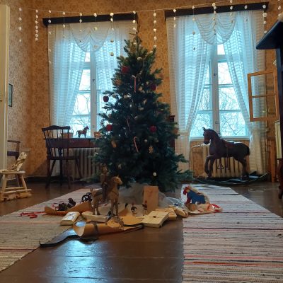 Lastenhuoneessa koristeltu joulukuusi, jonka juurella on vanhoja puisia leluja. Lattialla räsymatot. Katosta roikkuu valkoisia pumpulista tehtyjä joulukoristeita.
