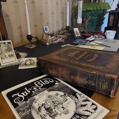 Etualalla kirjoituspöydällä lehti "Jul Blad" sekä vanha Raamattu "Det Heliga Skrift". Pöydällä kaksi kynttilää ja joulukorttien kirjoittaminen on kesken.