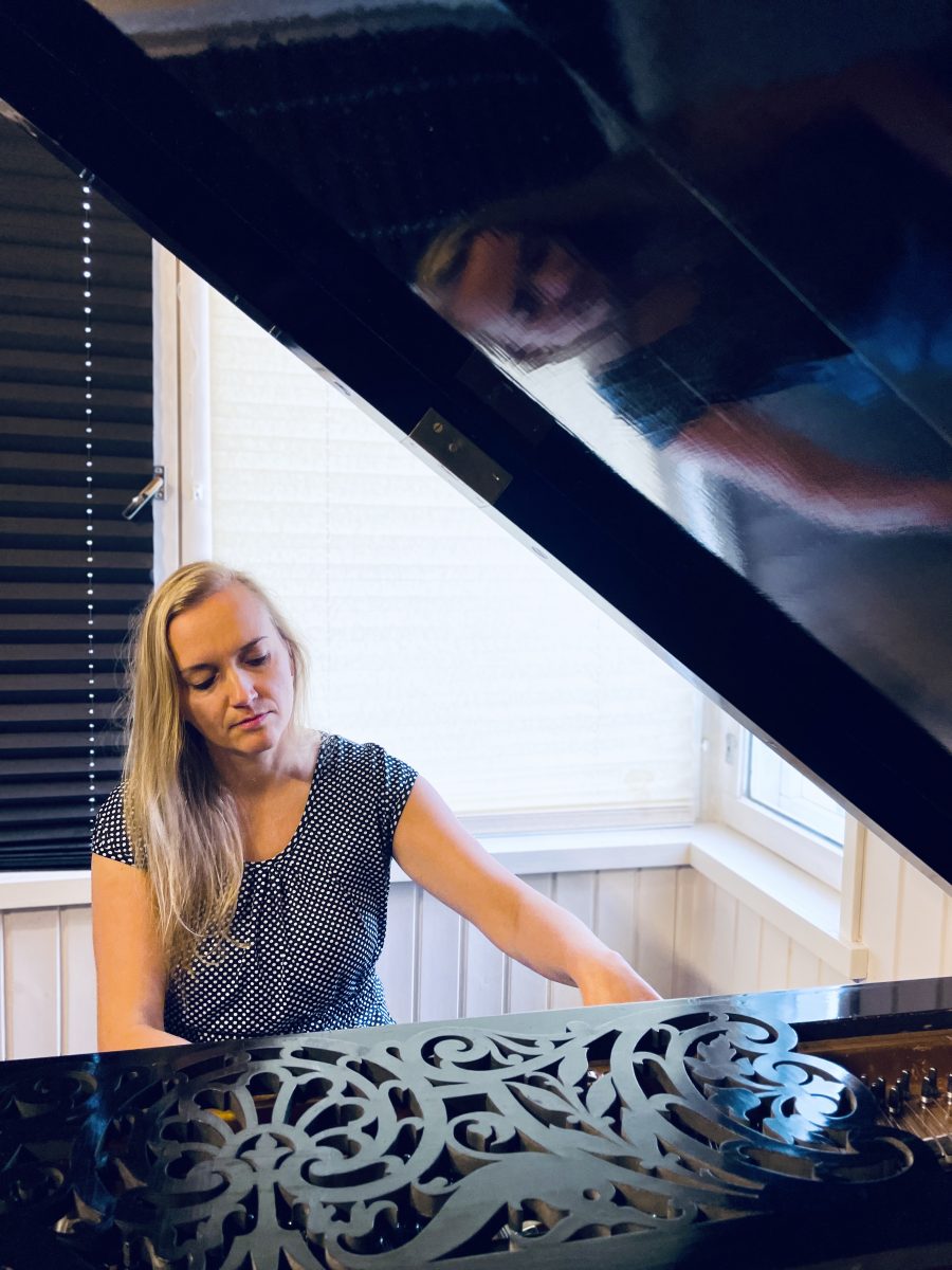 Flyygelin takaa otetussa kuvassa vaalea pitkätukkainen nainen lyhyhihaisessa paidassa soittaa pianoa. Flyygelin kansi on auki ja kuva rajautuu oikeasta kulmasta flyygelin kanteen.